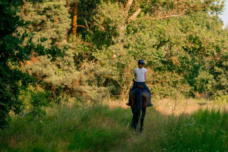 Téléchargez les photos : Une cavalière vêtue d'un casque monte son beau cheval noir dans la forêt lors d'une balade à cheval - en image libre de droit