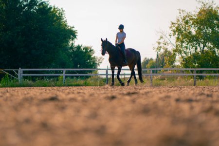 Foto de Una jinete vestida con un casco monta su hermoso caballo negro en una arena de equitación durante el paseo a caballo - Imagen libre de derechos