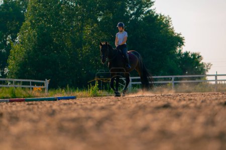 Foto de Una jinete vestida con un casco monta su hermoso caballo negro en una arena de equitación durante el paseo a caballo - Imagen libre de derechos