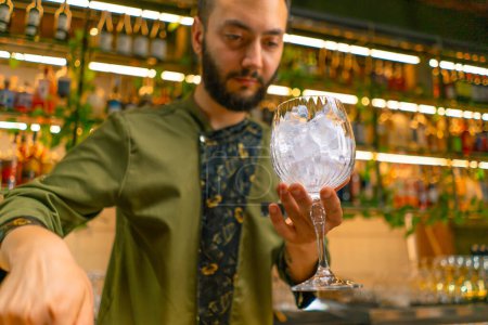 Téléchargez les photos : Barman professionnel tient un verre avec de la glace dans ses mains avant de faire un cocktail au bar - en image libre de droit