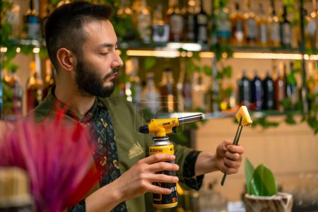 Téléchargez les photos : Un barman dans un bar ou un restaurant brûle un morceau d'ananas au feu décorer un cocktail alcoolisé - en image libre de droit