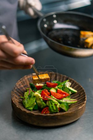 Photo for Professional kitchen in a chef restaurant puts ready-made salad with fried tofu cheese on plate close-up - Royalty Free Image