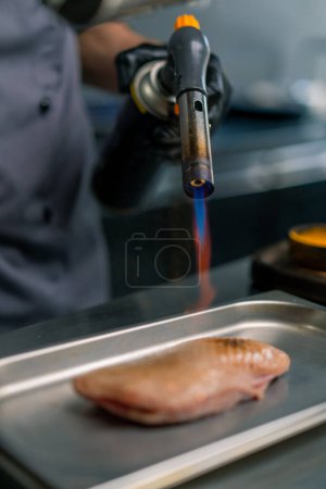 Téléchargez les photos : Gros plan main du chef avec brûleur à gaz friture poitrine de canard cru dans la cuisine professionnelle du restaurant - en image libre de droit