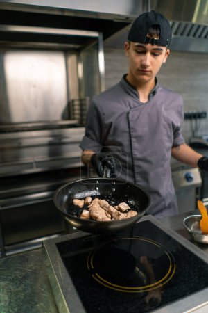 Foto de Retrato de un joven chef reflexivo en una cocina profesional en un restaurante freír filete de pollo en la sartén - Imagen libre de derechos