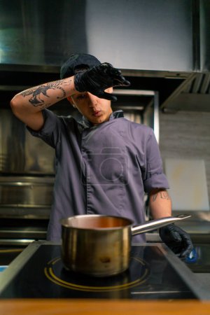 Foto de Chef cansado se limpia la frente con la mano mientras cocina la estufa de alimentos en olla en la cocina del restaurante - Imagen libre de derechos