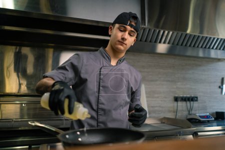 Photo for Portrait of a young thoughtful chef in a professional kitchen in a restaurant pouring oil into pan before cooking - Royalty Free Image