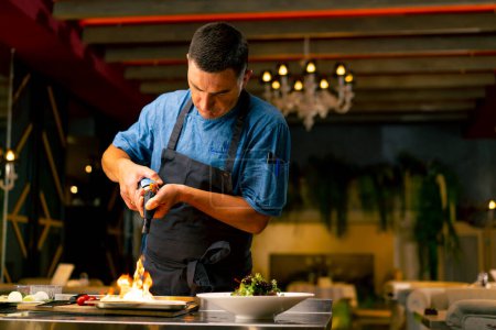 Foto de El chef de un restaurante italiano utiliza un quemador de gas para freír ingredientes mientras prepara una ensalada en la cocina profesional - Imagen libre de derechos