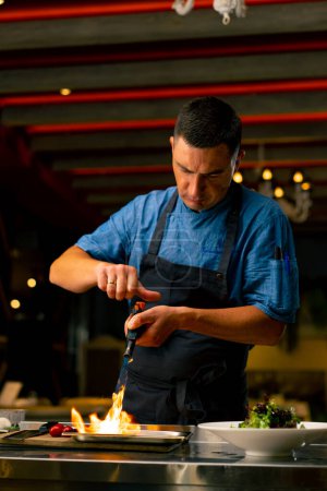 Foto de El chef de un restaurante italiano utiliza un quemador de gas para freír ingredientes mientras prepara una ensalada en la cocina profesional - Imagen libre de derechos