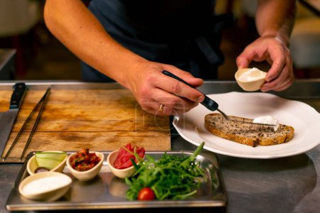 Foto de Un primer plano de un chef esparciendo queso crema sobre pan mientras prepara bruschetta en una cocina profesional en un restaurante italiano - Imagen libre de derechos