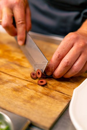 Foto de Primer plano de un chef cortando aceitunas con un cuchillo mientras prepara una ensalada en la cocina del restaurante italiano - Imagen libre de derechos