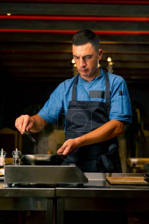 Photo for A close-up of a chef with tongs in his hand frying bacon for a burger in the professional kitchen of  Italian restaurant - Royalty Free Image