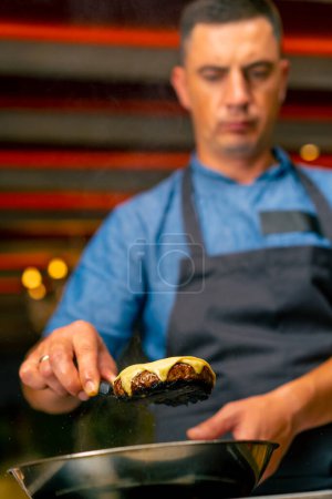 Téléchargez les photos : Gros plan d'un chef tenant une spatule avec laquelle il a enlevé une galette de fromage lors de la cuisson d'un hamburger dans la cuisine du restaurant - en image libre de droit