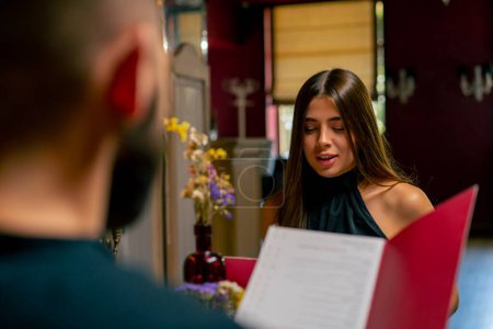 Téléchargez les photos : Jeune couple mec et fille assis dans un restaurant italien en regardant le menu et en choisissant ce qu'il faut commander pour le dîner pendant la date - en image libre de droit