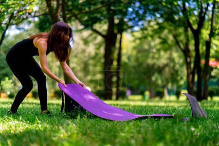 Téléchargez les photos : La fille pose un tapis de fitness sur l'herbe dans le parc pour commencer l'entraînement de méditation de yoga - en image libre de droit
