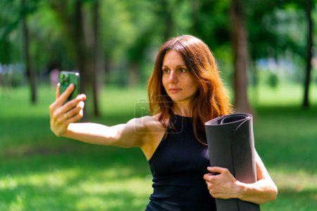 Téléchargez les photos : Active belle femme yogi lors d'une promenade dans le parc avant ou après l'entraînement prend une photo sur le téléphone tient tapis de fitness dans ses mains - en image libre de droit