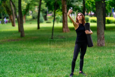 Photo for Active beautiful yogi woman during a walk in the park before or after training takes a photo on the phone holds fitness mat in her hands - Royalty Free Image