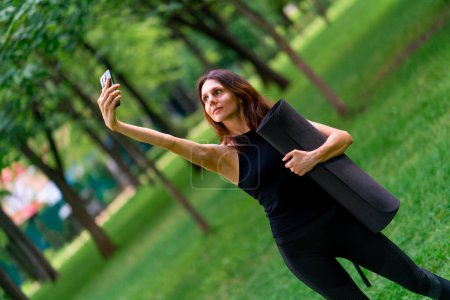 Téléchargez les photos : Active belle femme yogi lors d'une promenade dans le parc avant ou après l'entraînement prend une photo sur le téléphone tient tapis de fitness dans ses mains - en image libre de droit