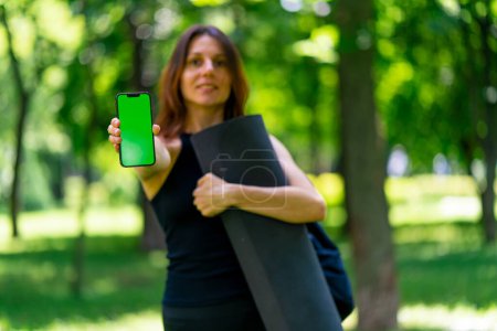 Foto de Mujer yogui hermosa activa durante un paseo por el parque antes o después del entrenamiento se para con una esterilla de fitness con un smartphone en las manos - Imagen libre de derechos