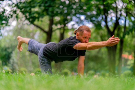 Foto de Hombre barbudo guapo meditando en el parque practicando yoga haciendo estiramientos y ejercicios diferentes grupos musculares - Imagen libre de derechos