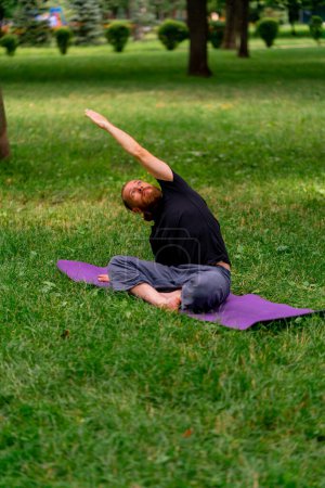 Foto de Hombre barbudo guapo meditando en el parque practicando yoga haciendo estiramientos y ejercicios diferentes grupos musculares - Imagen libre de derechos