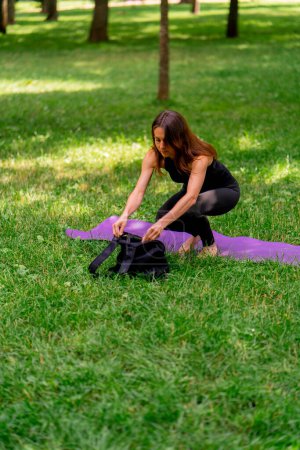 Téléchargez les photos : La jeune fille pose un tapis de fitness sur l'herbe dans le parc pour commencer à pratiquer la méditation de yoga sort l'équipement du sac - en image libre de droit