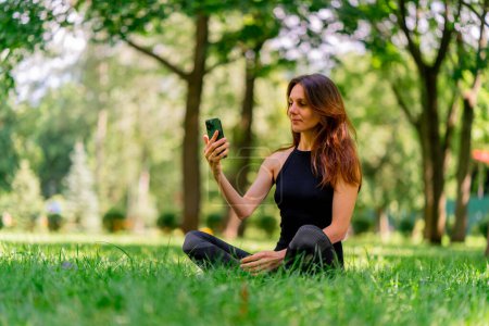 Photo for Active beautiful yogi woman during a walk in the park before or after training stands with a fitness mat holding smartphone in her hands - Royalty Free Image