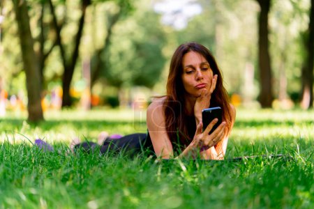 Téléchargez les photos : Active belle femme yogi se reposant dans le parc avant ou après l'entraînement parler via smartphone appel vidéo tout en étant couché sur tapis de fitness - en image libre de droit