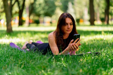Photo for Active beautiful yogi woman resting in the park before or after training talking via smartphone video call while lying on fitness mat - Royalty Free Image