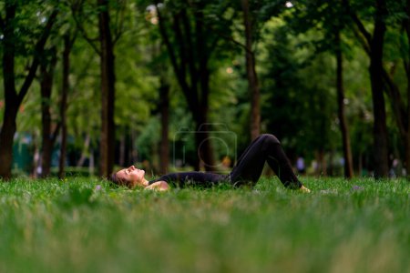 Téléchargez les photos : Active belle femme yogi reposant dans le parc de la ville après l'entraînement couché sur un tapis de fitness en regardant le ciel - en image libre de droit