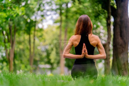 Photo for Experienced female instructor meditating in the park practicing yoga doing stretching and exercises different muscle groups back view - Royalty Free Image