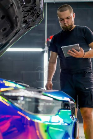 Photo for Car service worker is looking at a tablet in his hand while standing next to an expensive car with the hood open - Royalty Free Image