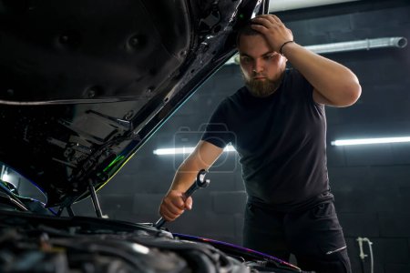 Foto de Un trabajador de servicio de coches con una llave en la mano está sosteniendo su cabeza y tratando de arreglar el coche con la campana abierta - Imagen libre de derechos