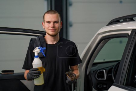 Téléchargez les photos : Portrait d'un jeune ouvrier de lavage de voiture tenant un pistolet pulvérisateur et une brosse sur le fond d'un VUS blanc en cours de démêlage - en image libre de droit