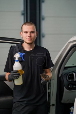 Foto de Retrato de un joven trabajador de lavado de coches sosteniendo una pistola de pulverización y cepillo contra el fondo de un SUV blanco en proceso de desenredar - Imagen libre de derechos