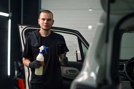 Photo for Portrait of a young car wash worker holding a spray gun and showing a thumbs up sign on the background of a white SUV in t process of detangling - Royalty Free Image