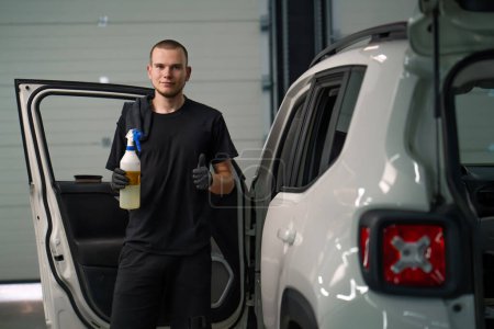 Photo for Portrait of a young car wash worker holding a spray gun and showing a thumbs up sign on the background of a white SUV in t process of detangling - Royalty Free Image