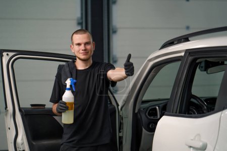 Foto de Retrato de un joven trabajador de lavado de coches sosteniendo una pistola de pulverización y mostrando un cartel de pulgares hacia arriba en el fondo de un SUV blanco en t proceso de desenredar - Imagen libre de derechos