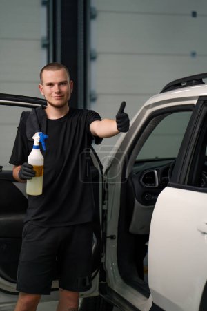 Foto de Retrato de un joven trabajador de lavado de coches sosteniendo una pistola de pulverización y mostrando un cartel de pulgares hacia arriba en el fondo de un SUV blanco en t proceso de desenredar - Imagen libre de derechos