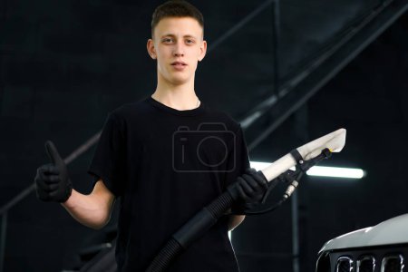 Téléchargez les photos : Portrait d'un jeune ouvrier de lavage de voiture tenant un aspirateur et montrant un pouce vers le haut signe sur le fond d'un 4x4 blanc en cours de démêlage - en image libre de droit
