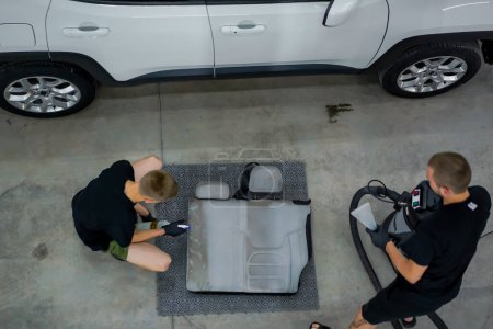 Téléchargez les photos : Deux employés de lavage de voiture retirent les sièges d'une voiture blanche et les nettoient à sec dans le processus de détailler auto - en image libre de droit