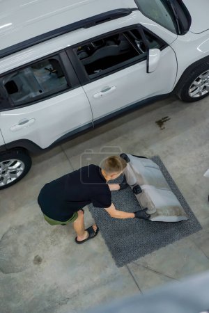 Foto de Dos empleados de lavado de coches quitan los asientos de un coche blanco y los limpian en seco en el proceso de detallar auto - Imagen libre de derechos