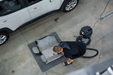 Téléchargez les photos : Un employé de lavage de voiture utilise un aspirateur pour nettoyer les sièges d'auto blancs en cours de démêlage vue de dessus - en image libre de droit