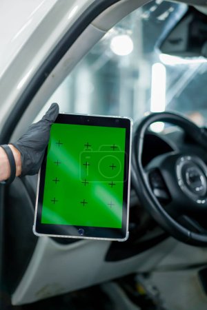 Téléchargez les photos : Gros plan d'une main tenant une tablette sur laquelle un écran vert est ouvert sur le fond de l'intérieur de la voiture - en image libre de droit