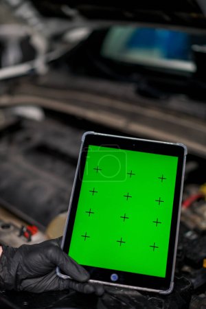 Foto de Primer plano de una mano con una tableta en la que la pantalla verde está abierta sobre el fondo del coche con la capucha abierta - Imagen libre de derechos
