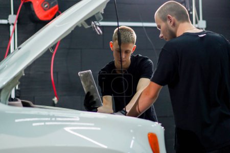 Foto de Dos mecánicos de automóviles están al lado de SUV blanco con la capucha abierta y mira la tableta que sostienen en sus manos mientras reparan el coche - Imagen libre de derechos