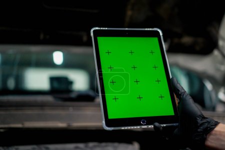 Téléchargez les photos : Gros plan d'une main avec une tablette sur laquelle l'écran vert est ouvert sur le fond de la voiture avec le capot ouvert - en image libre de droit
