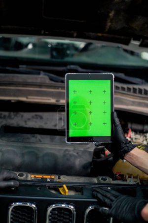Téléchargez les photos : Gros plan d'une main avec une tablette sur laquelle l'écran vert est ouvert sur le fond de la voiture avec le capot ouvert - en image libre de droit