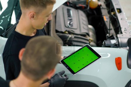 Téléchargez les photos : Deux mécaniciens automobiles se tiennent à côté du VUS blanc avec le capot ouvert et regardent une tablette avec un écran vert ouvert tout en réparant la voiture - en image libre de droit