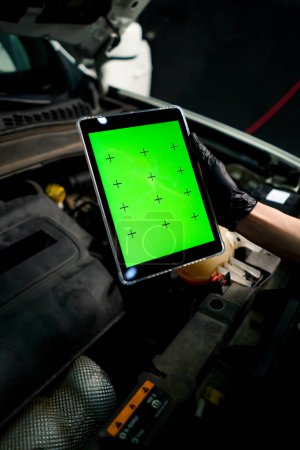 Photo for Close-up of a hand with a tablet on which the green screen is open against the background of car with the hood open - Royalty Free Image