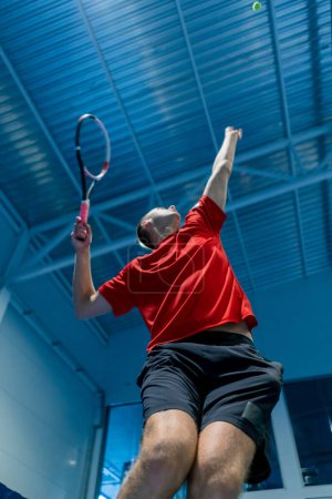 Foto de Joven en una cancha de tenis cubierta golpeando la pelota con una raqueta en un salto servir durante el juego de tenis instructor deporte profesional - Imagen libre de derechos